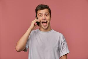 Indoor shot of overjoyed brown eyed young man in grey t-shirt posing over pink background, having excited conversation on phone and looking to camera cheerfully photo
