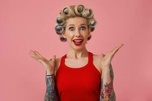 Close-up of amazed young attractive blonde woman with tattooes and evening makeup making hairdo and looking at camera with wide eyes opened, raising palms surprisedly over pink background photo