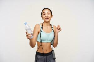 Cheerful young brown-eyed woman without makeup looking happily at camera while finishing her training, standing over white background with bottle of water in her hand photo