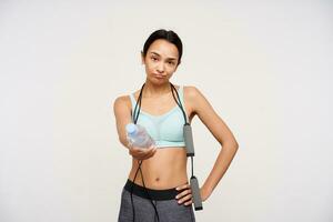 Displeased young brown-eyed slim brunette woman with jumping rope on her neck sharing with someone bottle on water while finishing her training, isolated over white background photo