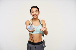 Active young positive brunette female smiling cheerfully while holding bottle of water, wearing jumping rope on her neck while standing over white background photo