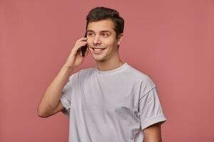 retrato de hermoso joven marrón ojos hombre en gris camiseta mirando aparte con encantador sincero sonrisa, participación móvil teléfono en mano y teniendo agradable conversación, en pie terminado rosado antecedentes foto