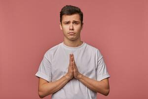 Studio shot of pretty dark haired young male with short haircut posing over pink background with raised hands, keeping palms together and begging for something pitifully photo