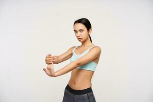 Indoor shot of attractive young brown haired woman pulling her fingers while stretching muscules and looking attentively at camera, isolated over white background photo