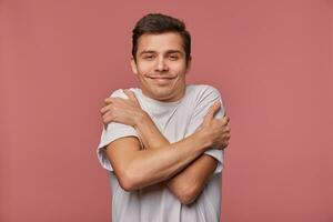 Indoor portrait of cute dark haired young man looking to camera with sincere wide smile and hugging himself, standing over pink background in grey t-shirt photo