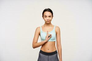 Bewildered young pretty brunette woman dressed in sporty clothes raising wonderingly her eyebrow while looking at camera and showing namaste sign, isolated over white background photo