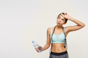 Exhausted young brown haired woman with ponytail hairstyle wiping the sweat with raised hand after hard training, holding water while posing over white background photo