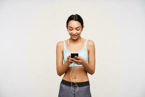 Good looking young sporty brown haired woman dressed in mint sporty bra and grey leggins holding mobile phone and looking gladly on screen, posing over white background photo