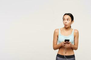 Surprised young slim brunette woman with ponytail hairstyle looking wonderingly aside while holding mobile phone, isolated over white background in sporty clothes photo