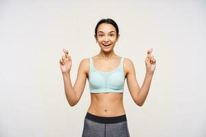 Excited young attractive slim brown haired lady looking emotionally at camera and raising hands with crossed fingers, standing against white background photo