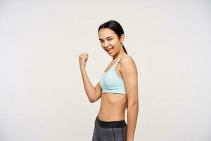 Indoor photo of young pretty brown haired slim lady raising happily fist in yes gesture while standing over white background in mint sporty bra and grey leggins