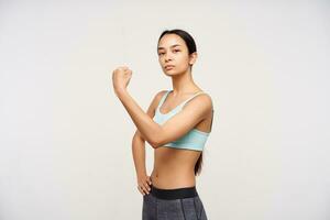 Indoor photo of young attractive sporty brown haired woman demonstrating her strong hand and looking proudly at camera, standing over white background