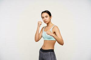 Severe young attractive brown haired sporty woman keeping her lips folded while standing white background with raised fists. Sport and healthy lifestyle concept photo