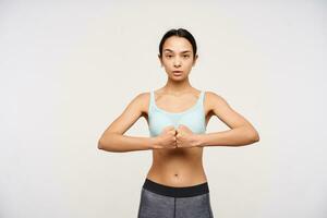 Studio photo of young brown haired athletic lady folding her fists together while looking menacingly at camera, standing over white background in sporty clothes