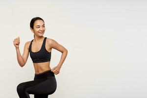 Studio shot of glad slim young brown haired female lady with casual hairstyle being in nice mood while making morning workout, standing over white background photo