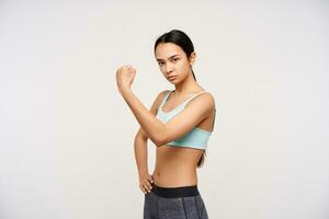 Serious young brown-eyed long haired brunette woman raising hand while showing strong biceps and looking severely at camera, isolated over white background photo