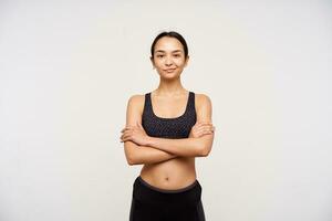 Studio photo of young slim brown haired woman in sporty wear folding her hands on chest while looking positively at camera with gentle smile, standing over white background