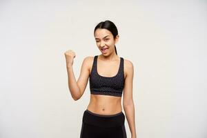 Indoor photo of young pretty brown haired woman keeping one eye closed while looking happily at camera with raised hand, standing over white background