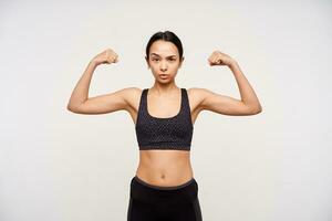 Severe young sporty brown haired woman raising her hands while demonstrating strong biceps and looking seriously at camera, isolated over white background photo