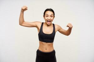 Agitated young lovely dark haired woman in black sporty wear raising emotionally her hands while looking excitedly at camera with wide mouth opened, posing over white background photo