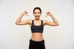 Studio shot of young attractive brunette female with ponytail hairstyle thumbing joyfully on herself while looking excitedly at camera, isolated over white background photo