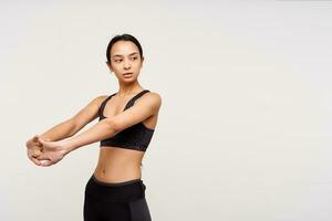 Serious young slim pretty brown haired woman with casual hairstyle training her arms while standing over white background and looking pensively aside photo