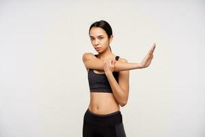Studio shot of young sporty dark haired woman in sporty clothes stretching her arms and looking seriously at camera, isolated over white background in athletic wear photo