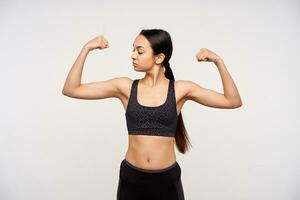 Indoor shot of young long haired brunette sporty lady looking attentively on her raised hand while standing over white background in black top and leggins photo