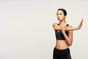 Indoor photo of young brown haired slim woman looking ahead with concentrated face while stretching her muscules after training, standing over white background