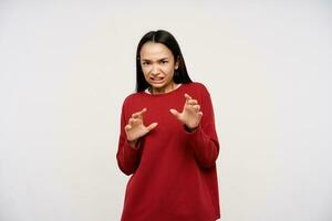 Young lady, afraid asian woman with dark long hair. Wearing red sweater and trying to protect herself with hands. Watching scared at the camera isolated over white background photo