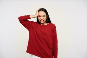 Bewildered young dark haired brunette lady with natural makeup keeping raised hand on her head and grimacing face while looking at camera, isolated over white background photo