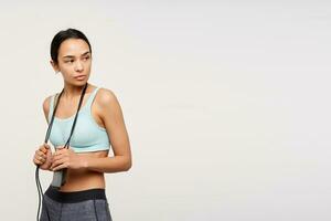 Young sporty lady, serious asian woman with dark long hair. Wearing sportswear and holding a jump rope over her neck. Watching to the left at copy space, isolated over white background photo