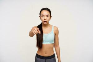Teenage girl, shocked looking asian woman. With black hair gathered in a ponytail, wearing sportswear. Watching and pointing at the camera, can not believe her eyes, isolated over white background photo