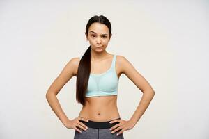 Teenage sporty girl, suspicious looking asian woman with dark long hair. Wearing sportswear and holding hands on a waist. Watching at the camera with eyebrow lifted, isolated over white background photo