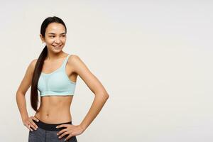 Portrait of attractive, sporty asian girl with dark long hair gathered in a ponytail. Wearing sportswear and holding hands on a waist. Watching to the right at copy space over white background photo