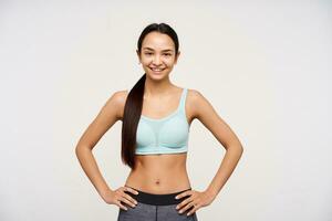 Young lady, sporty asian woman with dark long hair gathered in a ponytail. Wearing sportswear and holding hands on a waist, smiling. Watching at the camera isolated over white background photo