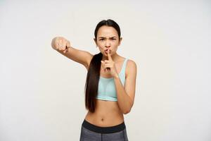 Portrait of serious, sporty, adult asian girl with dark long hair. Wearing sportswear and showing silence sign. Angry pointing and watching at the camera isolated over white background photo