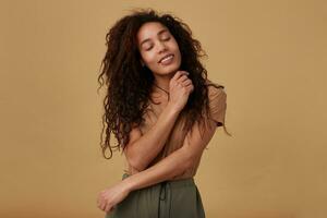 Goog looking young glad brown haired curly dark skinned woman keeping her eyes closed while smiling pleasantly and embracing her with raised hands, isolated over beige background photo