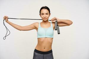Young lady, sporty asian woman with dark long hair. Wearing sportswear and stretching a jump rope over her neck. Watching confident at the camera isolated over white background photo