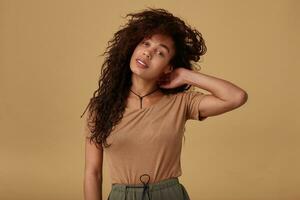 Attractive young dark skinned brunette woman with wild curly hair raising hand to her head and looking calmly at camera, standing over beige background photo