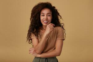 Happy young attractive long haired curly dark skinned female touching gently her face and looking joyfully at camera with broad smile, isolated over beige background photo