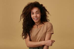 Joyful young attractive long haired curly dark skinned lady with casual hairstyle looking happily at camera with wide sincere smile while standing over beige background photo