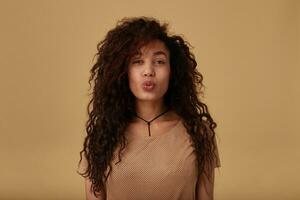 Studio photo of young lovely brown haired curly female with dark skin folding cheerfully her lips in air kiss while standing over beige background in nude t-shirt