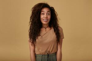 Agitated young lovely brown haired curly woman with dark skin raising surprisedly her eyebrows while looking positively at camera, isolated over beige background photo