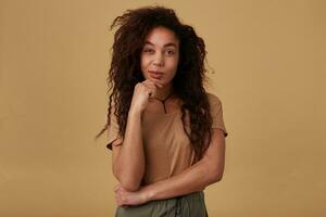 Indoor photo of young pretty brown haired curly dark skinned lady keeping raised hand on her face and looking dreamily at camera, isolated over beige background