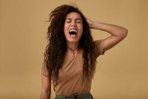 Pissed-off young long haired curly dark skinned female keeping her eyes closed while screaming emotionally and holding hand on her head, isolated over beige background photo