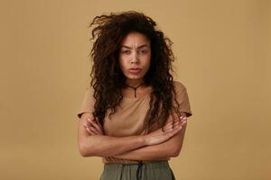 Offended young pretty curly dark skinned brunette lady folding her hands on chest while looking sadly at camera and frowning her eyebrows, posing over beige background photo