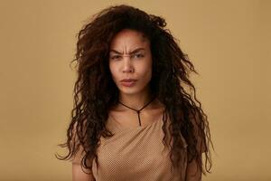 Gloomy young long haired curly dark skinned woman frowning her eyebrows while looking seripusly at camera with folded lips, isolated over beige background photo