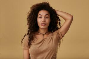 Portrait of charming young long haired curly dark skinned female with voluminous hairstyle raising hand to her head and looking calmly at camera, standing over beige background photo