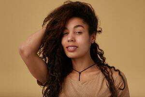 Portrait of young lovely curly dark skinned brunette lady with wild hairstyle looking gently at camera with soft smile while posing over beige background with raised hand photo